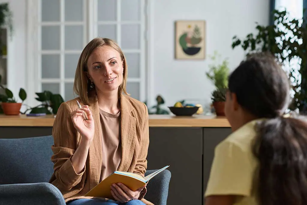 A female psychiatrist speaks to her patient, a teen girl sitting across from her, in her office