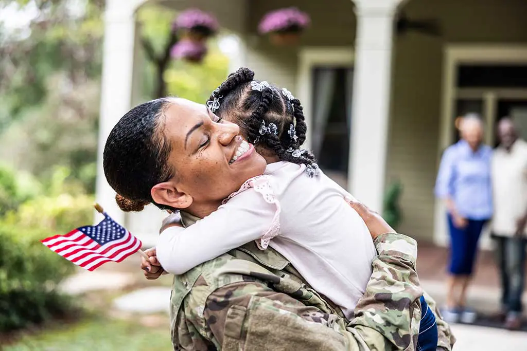 A mother in a military uniforms hugs her small daughter, who holds a little American flag