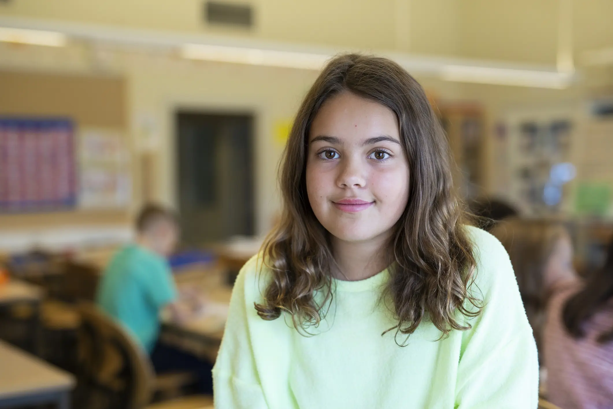 A pre-teen girl in a light green sweater smiles shyly while in a classroom