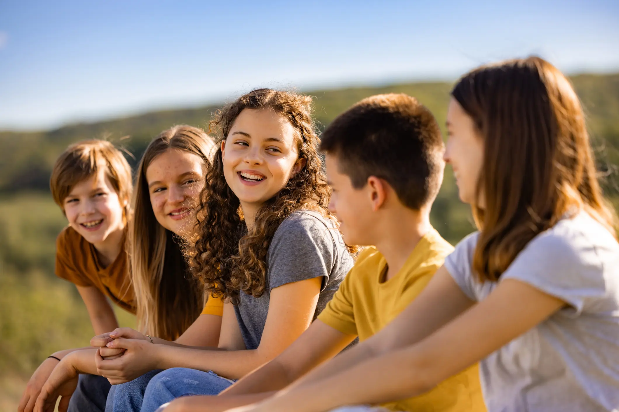 A group of pre-teens sit together in a field outdoors on a sunny day
