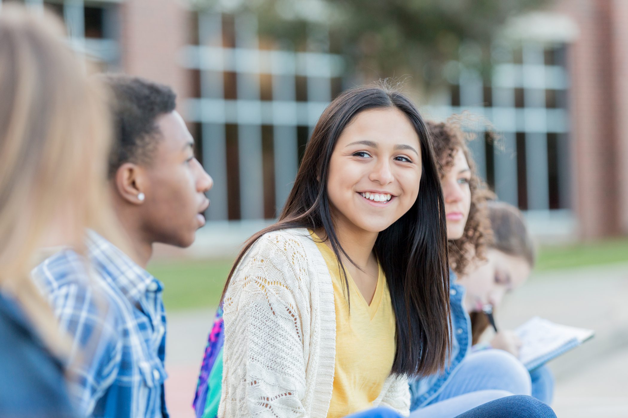 Teen girl smiling while sitting on a bench in front of school with her friends