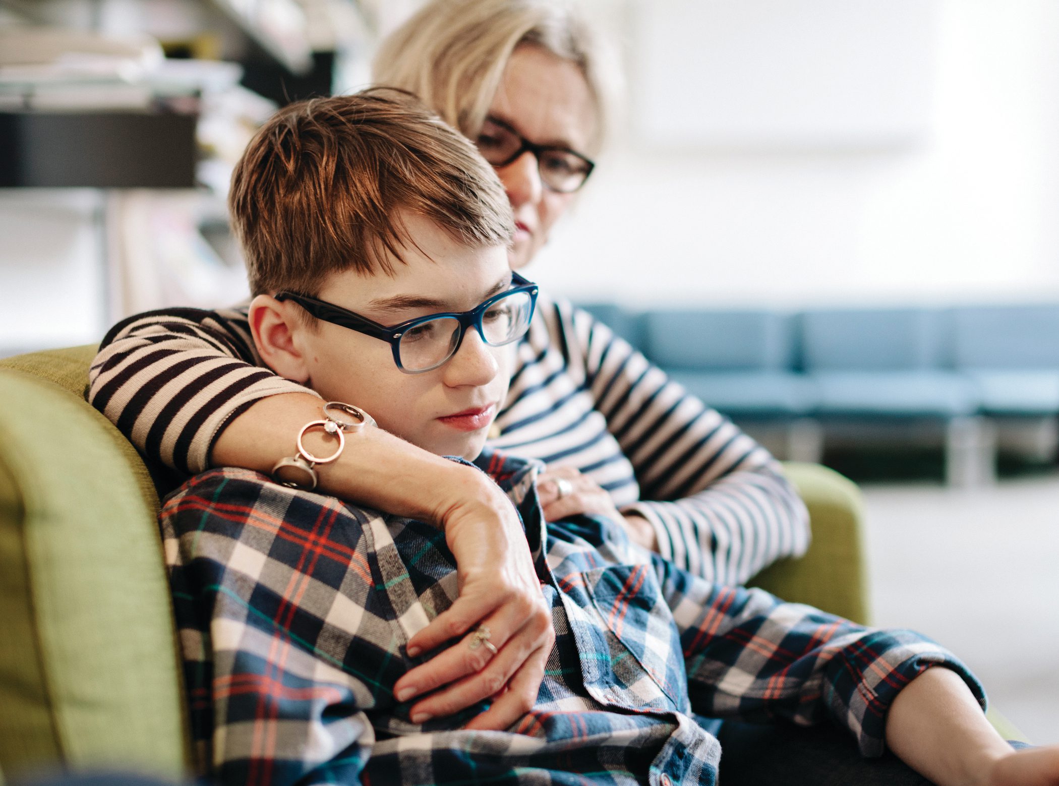 A mother wraps her arm around her sad son as they sit in a hospital lobby